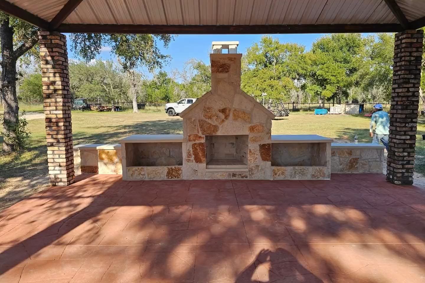 A finished outdoor pavilion with stone columns, a stone fireplace and stamped concrete under a metal roof
