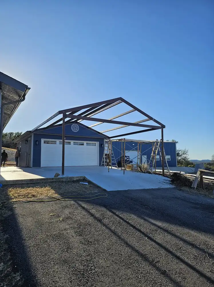 A bare steel carport truss standing next to a finished blue garage on new concrete